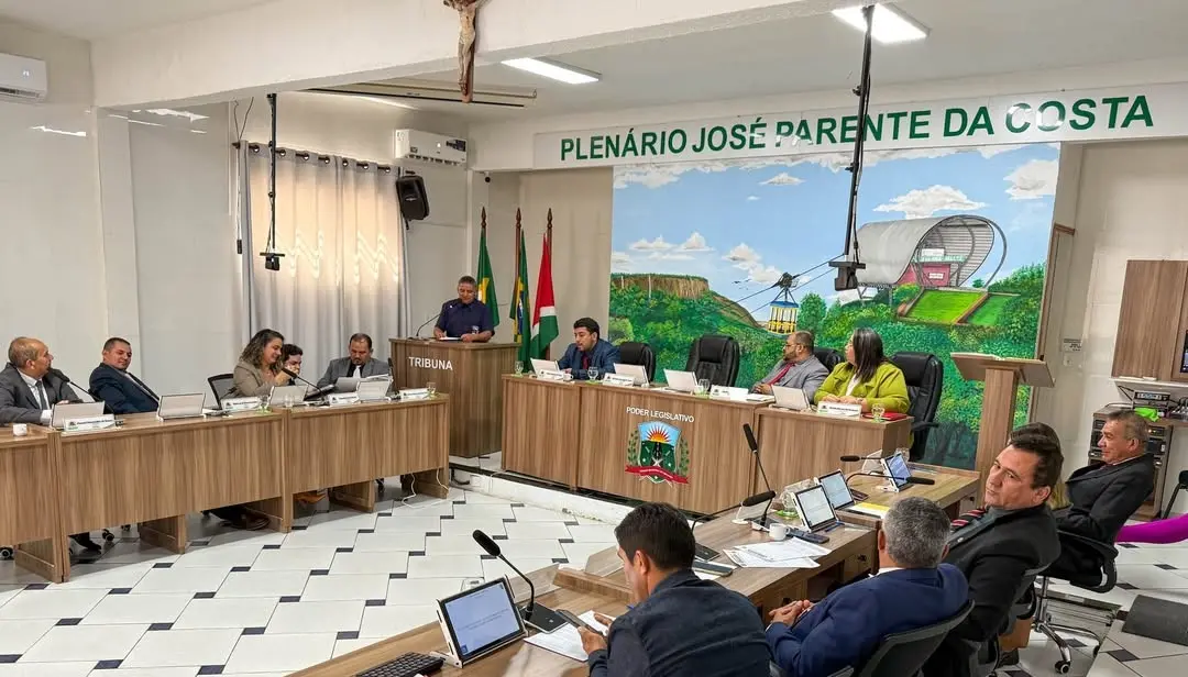 Sessão da Câmara Municipal em plenário com vereadores reunidos em torno de mesas em formato de U. Um homem fala na tribuna. Ao fundo, mural colorido com ilustração do teleférico de Ubajara e paredes com o nome ‘Plenário José Parente da Costa’. Vários vereadores utilizam notebooks durante a sessão.