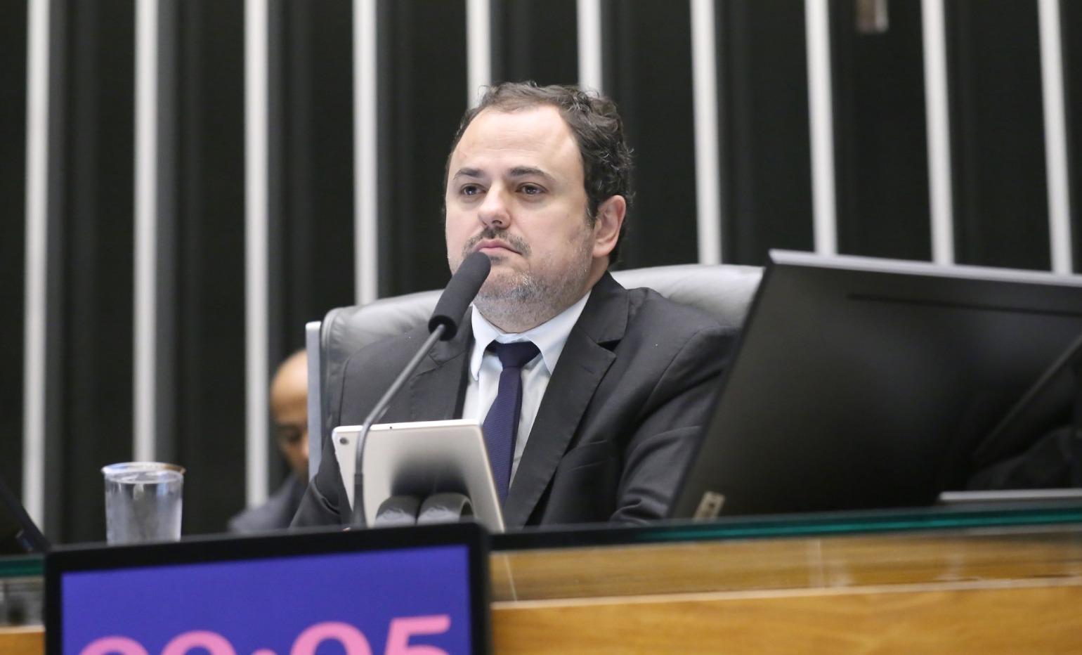 O deputado Glauber Braga é um homem de meia-idade, branco e de barba rala, grisalha e cabelo preto. Na foto, ele está sério e usa terno cinza com camisa social branca e gravata azul.