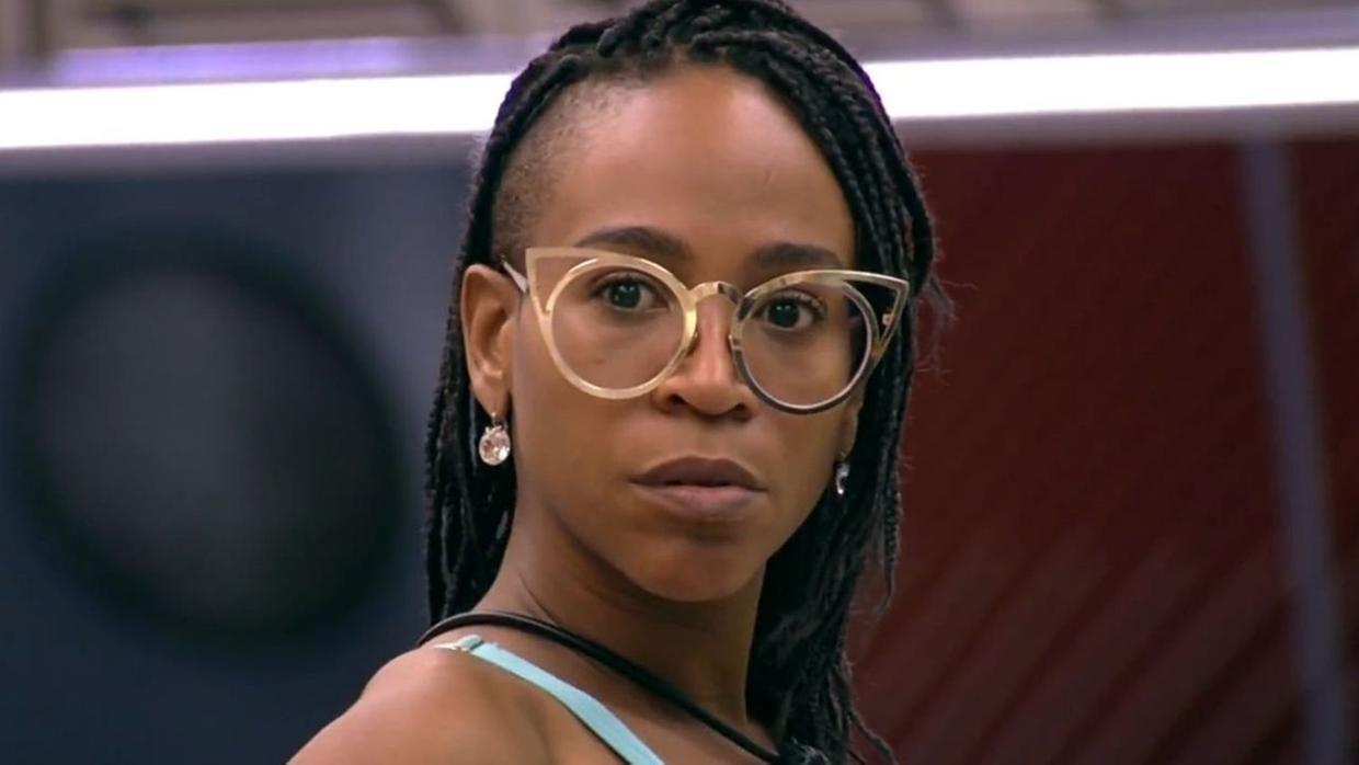 Karol é uma mulher jovem, negra e que usa óculos de grau redondos e tranças no cabelo. Na foto, ela está séria.