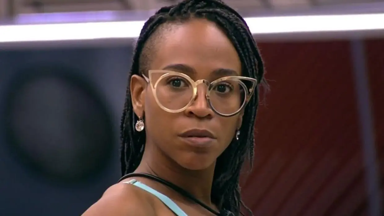 Karol é uma mulher jovem, negra e que usa óculos de grau redondos e tranças no cabelo. Na foto, ela está séria.