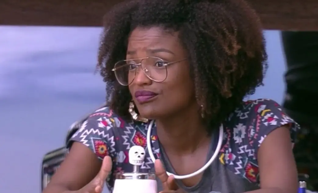 Nayara é uma mulher negra, jovem, que usa óculos de grau redondos e finos. Na foto, ela está com a expressão séria.