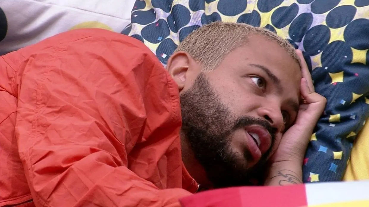 Projota é um homem negro de pele clara e cabelo curto loiro e barba preta. Na foto, ele está deitado de lado, dialogando com alguém.