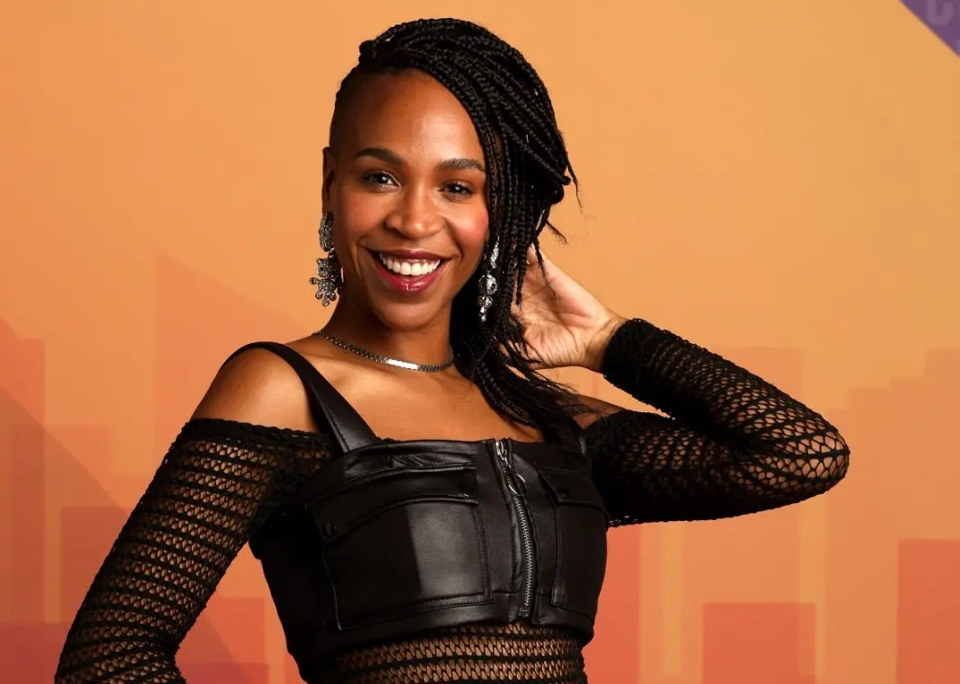 Karol é uma mulher jovem negra de cabelo trançado. Na foto, ela sorri enquanto posa.