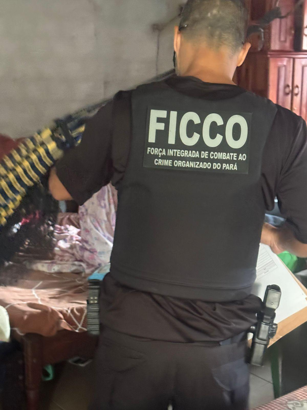 Agente da FICCO (Força Integrada de Combate ao Crime Organizado) do Pará visto de costas durante uma operação, vestindo colete tático e com arma na cintura, em ambiente interno.