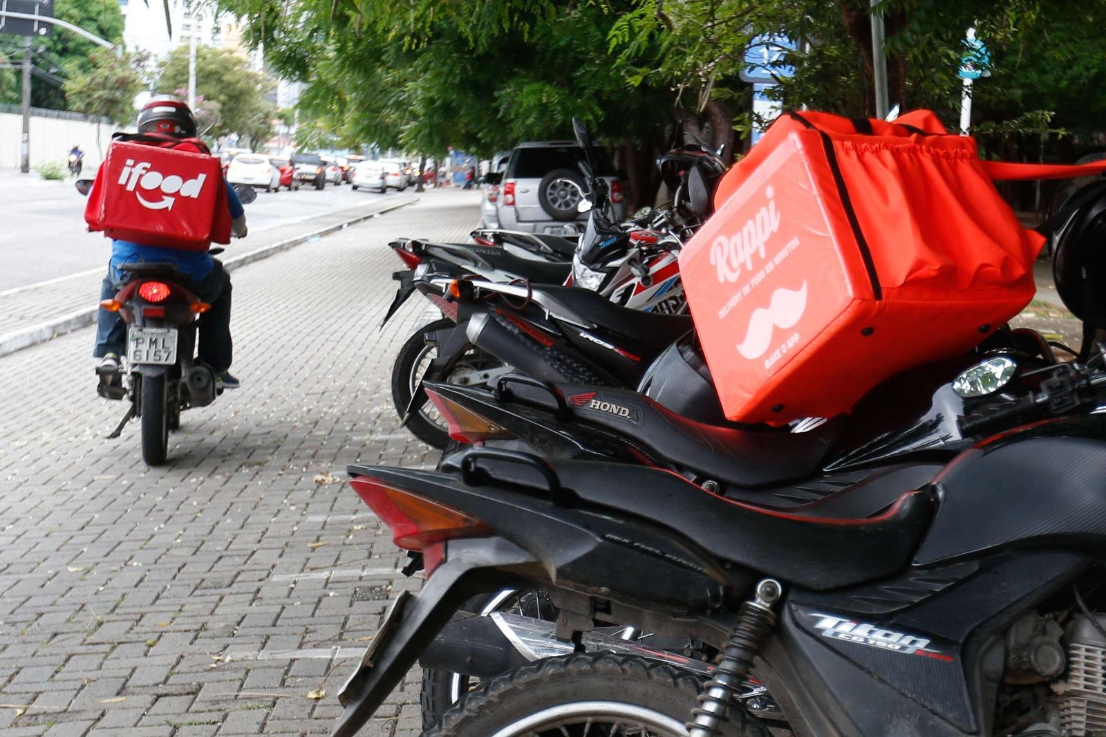 Imagem mostra motociclista, de costas, pilotando moto, levando mochila de aplicativo de entrega Ifood. Há outras motos estacionadas, um delas também tem uma mochila de entrega por aplicativo.