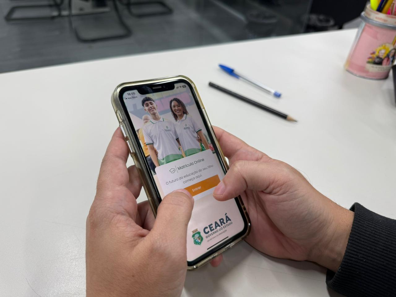 Mãos seguram um celular exibindo a tela inicial do aplicativo Matrícula Online do Governo do Ceará, com a imagem de dois estudantes sorrindo.
