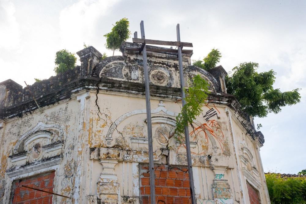 Fachada de prédio antigo e abandonado, com estrutura deteriorada, rachaduras visíveis na parede, janelas vedadas por tijolos e vegetação crescendo no telhado.