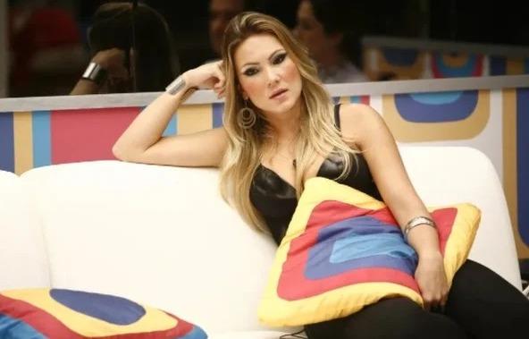 Josy é uma mulher jovem, branca, loira e de cabelo longo e liso. Na foto, ela está no sofá da casa do BBB.