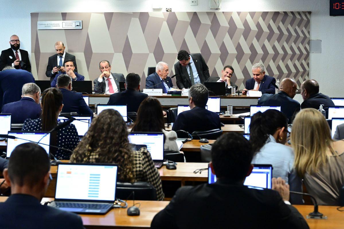 Sessão em comissão parlamentar com diversos participantes sentados diante de notebooks, acompanhando apresentação e discussões em ambiente institucional com painel geométrico ao fundo.