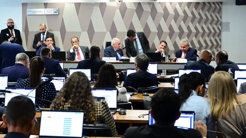 Sessão em comissão parlamentar com diversos participantes sentados diante de notebooks, acompanhando apresentação e discussões em ambiente institucional com painel geométrico ao fundo.