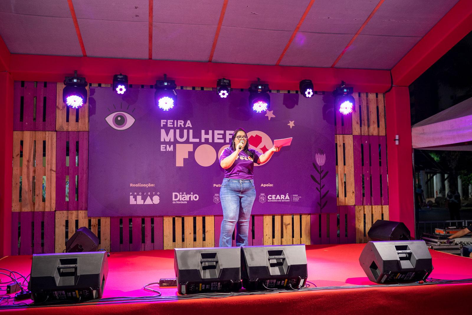 Mulher fala ao microfone no palco da Feira Mulher em Foco.