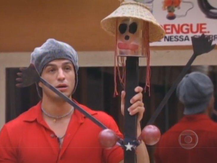 Participante do Big Brother Brasil segurando um boneco artesanal com chapéu de palha, olhos pintados e braços abertos, em cenário interno com parede bege e cartaz informativo ao fundo.