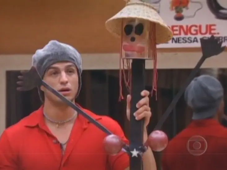 Participante do Big Brother Brasil segurando um boneco artesanal com chapéu de palha, olhos pintados e braços abertos, em cenário interno com parede bege e cartaz informativo ao fundo.