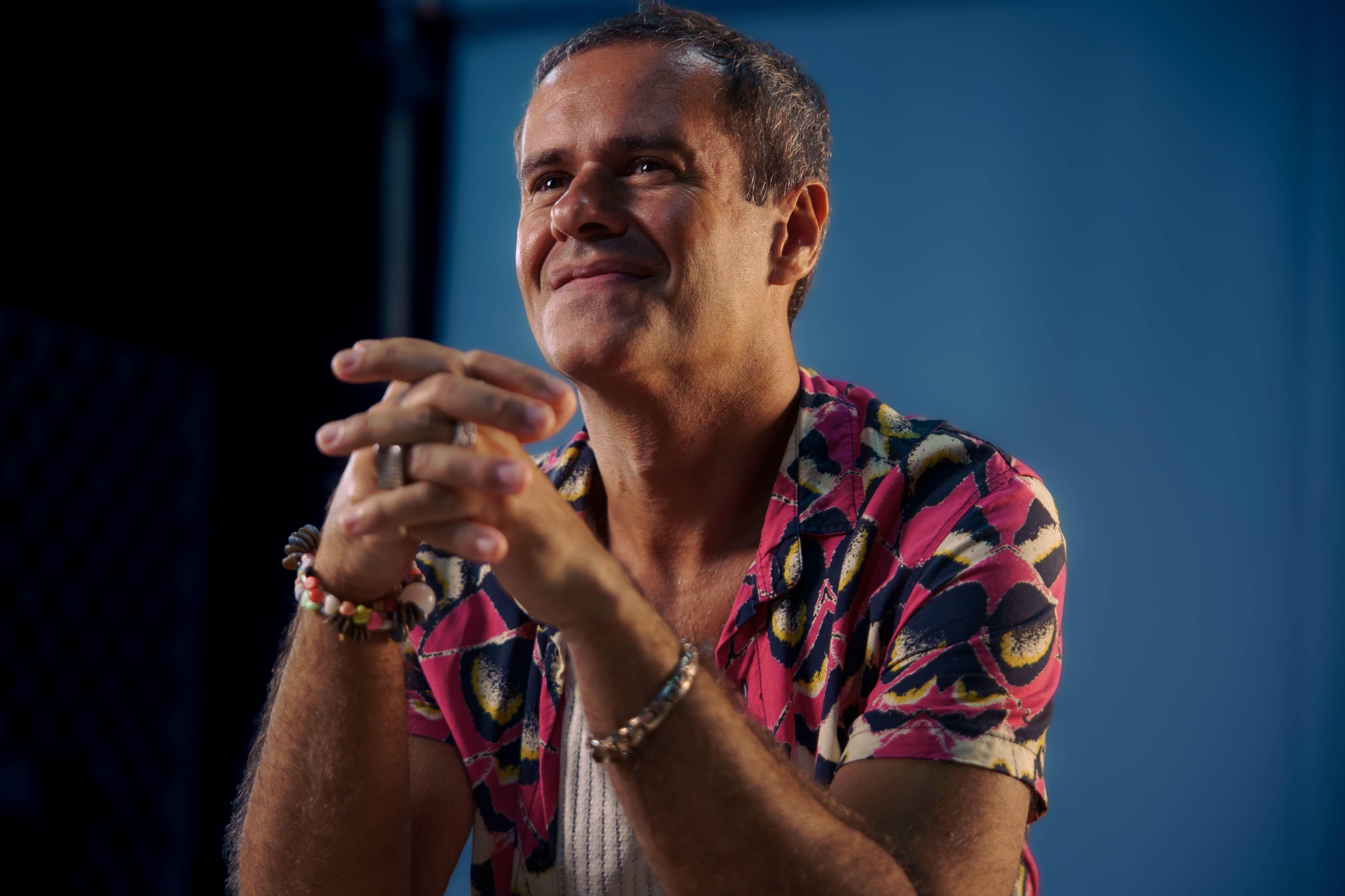 A imagem em plano médio e foco nítido captura Marcelo Jeneci, músico e cantor brasileiro. Ele está sentado, com o corpo ligeiramente inclinado para a frente, e parece estar em uma conversa ou apresentação, julgado pela iluminação de palco.
Jeneci tem a pele clara, cabelo curto e escuro, e está sorrindo de forma gentil, olhando para o canto superior esquerdo (fora da câmera). Suas mãos estão unidas à frente do peito, com os dedos entrelaçados em uma pose atenta e reflexiva.
Ele veste uma camisa de botão de manga curta com uma estampa vibrante e colorida, que combina tons de rosa choque, vermelho, preto e amarelo sobre uma base mais clara, remetendo a padrões tropicais ou de borboletas. Por baixo, ele usa uma camiseta regata branca. Em seu pulso esquerdo, há uma pilha de pulseiras de contas coloridas e, no pulso direito, algumas pulseiras em tons de terra e dourado.
O fundo é simples e escuro, com uma iluminação de cor azul turquesa suavemente iluminando a área atrás dele e criando um contraste que destaca a pessoa em primeiro plano. A iluminação geral é quente e dramática, focada no rosto e na parte superior do corpo do artista.