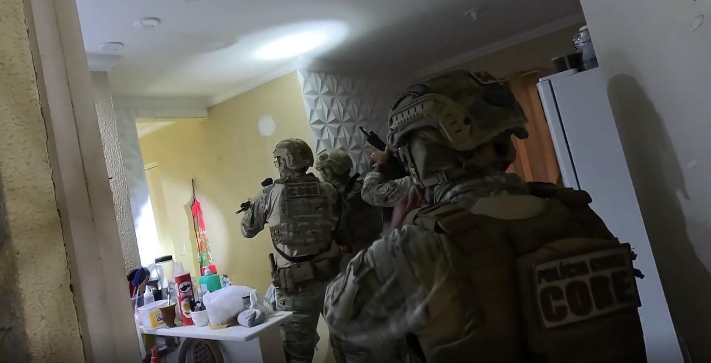 A foto mostra policiais civis, fardados, armados e com coletes balísticos, dentro de uma casa, durante uma operação policial.