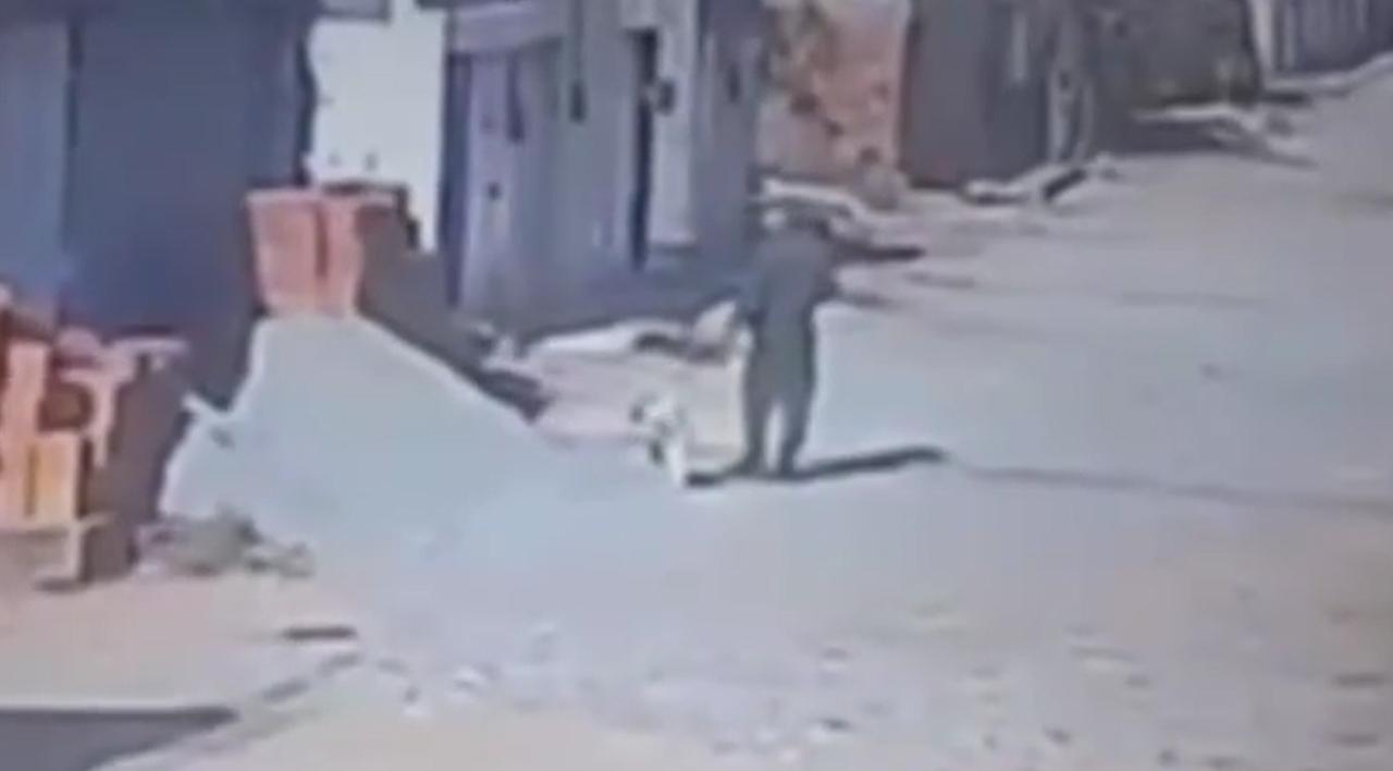 Imagem borrada de câmera de segurança mostrando silhueta de policial fardado levando um cachorro branco pequeno na coleira por uma rua deserta e com tijolos e areia de construção.