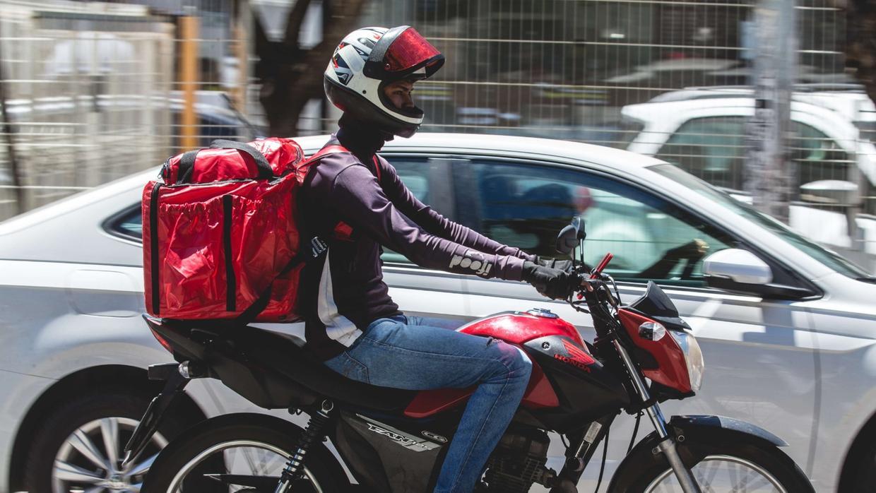 Motociclista circulando por Fortaleza vestindo calça jeans, camisa longa preta e com mochila de entrega vermelha nas costas.