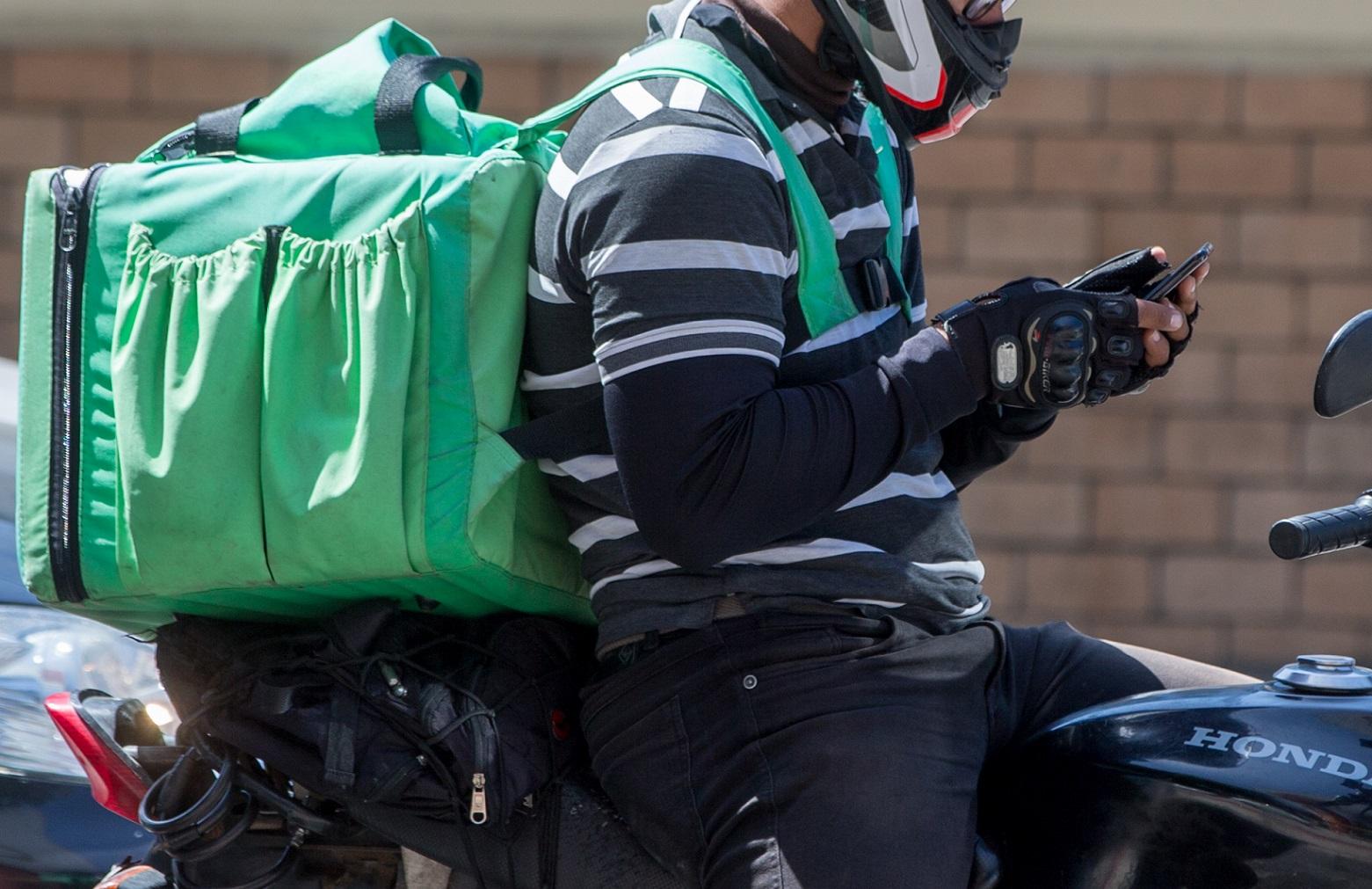 Entregador com capacete e mochila térmica verde nas costas, usando o telefone enquanto está parado na motocicleta.
