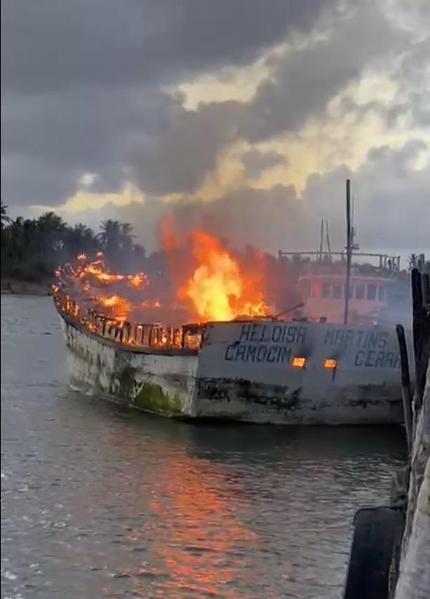 A imagem mostra um barco branco sendo incendiado, em alto mar. A embarcação tem escrito 'Heloísa Martins, Camocim Ceará'.