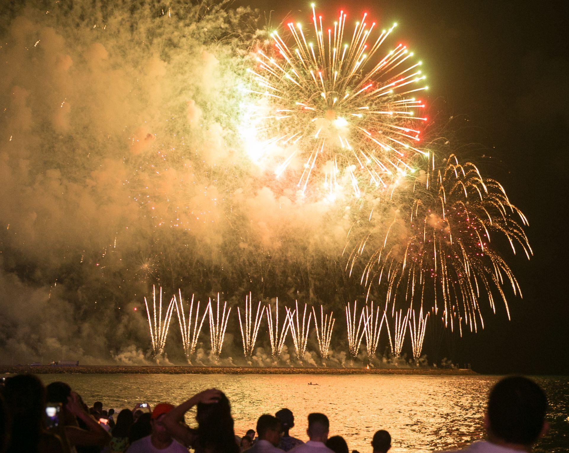 Show de fogos de artifício no céu noturno sobre o mar, com a silhueta de uma multidão assistindo.