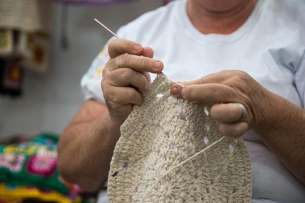 Na imagem, mãos de uma artesã mais velha segurando uma agulha de crochê rosa e um fio branco, trabalhando em um círculo ou peça de crochê em tom bege com um padrão rendado e texturizado. A artesã veste uma camiseta branca e usa um anel no dedo anelar direito.