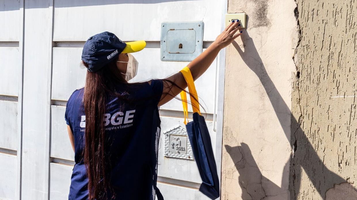 foto de mulher de cabelo longo liso e escuro, de costas e com máscara no rosto, tocando campainha de casa com portão branco. Ela usa blusa azul escura com nome IBGE e boné azul e amarelo.