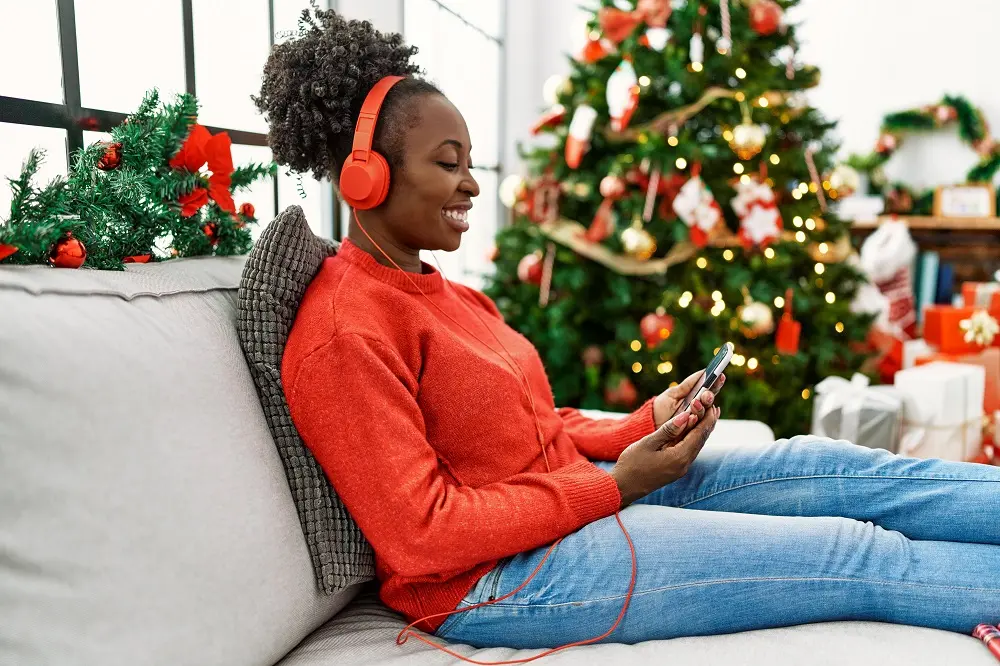 Uma mulher sorri enquanto ouve algo em um fone de ouvido e observa a tela de um celular. Ela é negra, tem cabelos pretos e cacheados presos num coque, usa suéter vermelho e calça jeans e está em frente a uma árvore de Natal.
