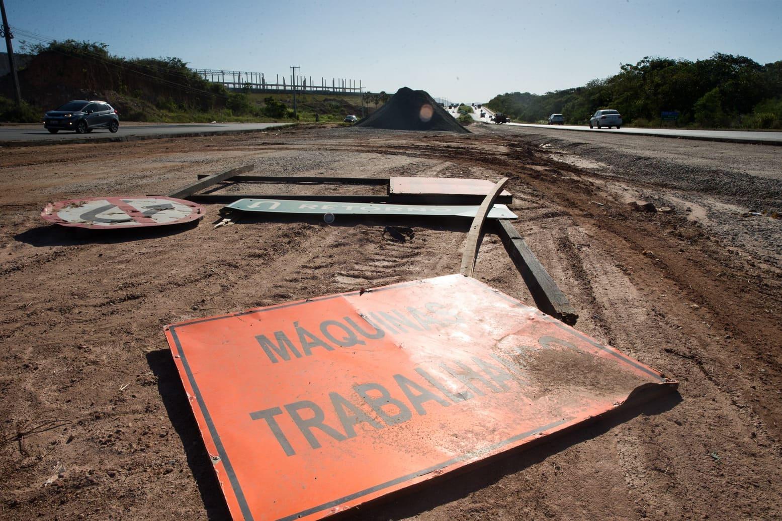 Placas de sinalização no Anel Viário caídas sobre o chão de terra em trecho de obra no Anel Viário; ao fundo, pista com carros passando e montes de brita usados na construção.