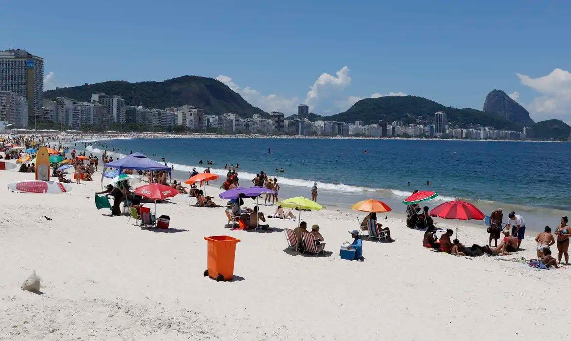 Foto mostra areia da praia de copacabana, com pessoas embaixo de guarda-sóis; ao fundo, está o mar e o pão-de-açúcar.