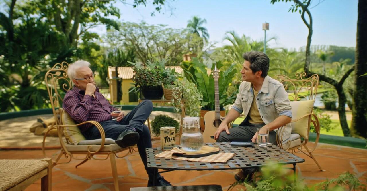 Moacyr e Daniel, respectivamente à esquerda e à direita, sentados em cadeiras de adornos dourados em um fundo de plantas. O Moacyr é um homem idoso de cabelos brancos, com óculos de grau preto e vestindo uma blusa de botão marrom e calça jeans. Daniel é um homem branco de meia idade, com cabelos lisos e pretos, e veste uma blusa amarela apagada, uma jaqueta branca desbotada e calça jeans.