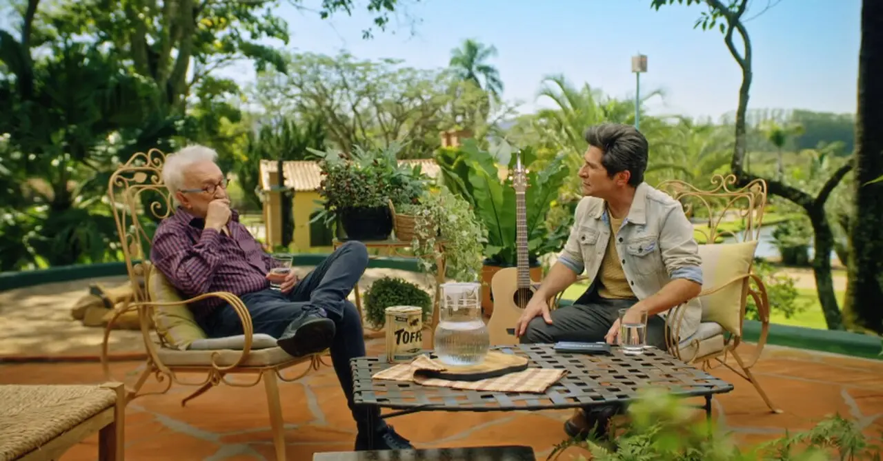 Moacyr e Daniel, respectivamente à esquerda e à direita, sentados em cadeiras de adornos dourados em um fundo de plantas. O Moacyr é um homem idoso de cabelos brancos, com óculos de grau preto e vestindo uma blusa de botão marrom e calça jeans. Daniel é um homem branco de meia idade, com cabelos lisos e pretos, e veste uma blusa amarela apagada, uma jaqueta branca desbotada e calça jeans.