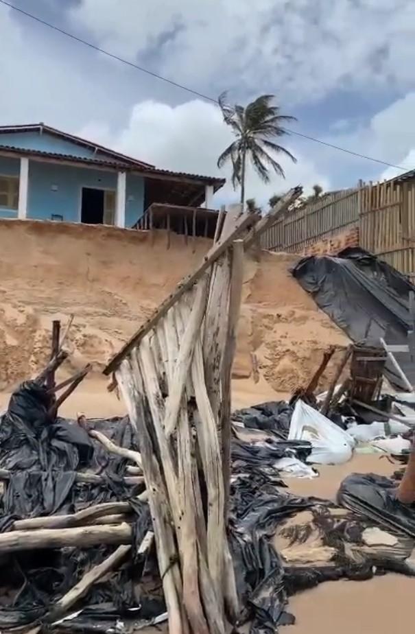 Praia da Peroba com sinais de forte erosão costeira, exibindo uma casa azul à beira de um barranco de areia íngreme e detritos de madeira e plástico espalhados na areia.
