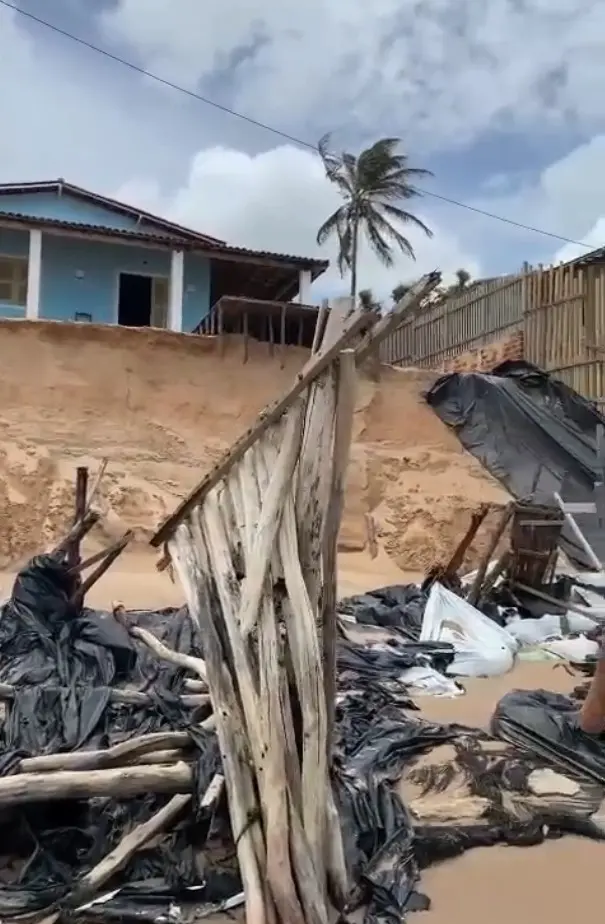 Praia da Peroba com sinais de forte erosão costeira, exibindo uma casa azul à beira de um barranco de areia íngreme e detritos de madeira e plástico espalhados na areia.