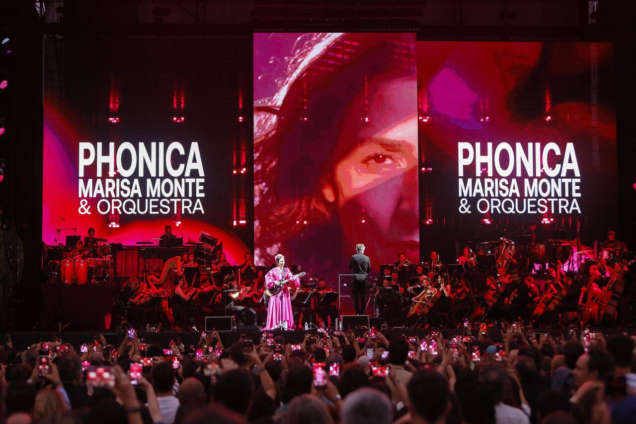 Mulher branca de vestido rosa em cima de palco com violão na mão. Ao redor, ambiente com luz vermelha e letreiro no palco com rosto da mulher e os dizeres 