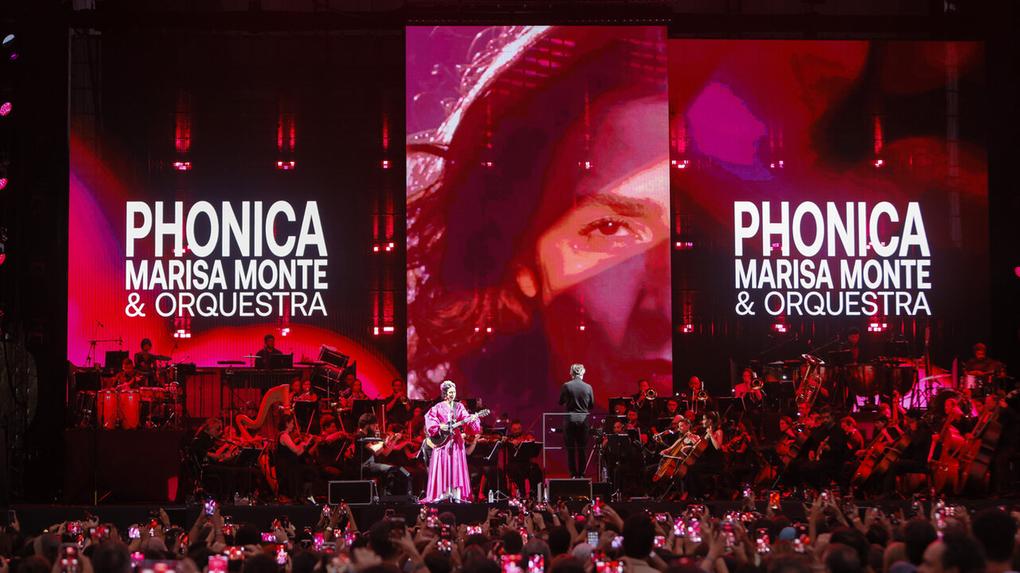 Mulher branca de vestido rosa em cima de palco com violão na mão. Ao redor, ambiente com luz vermelha e letreiro no palco com rosto da mulher e os dizeres