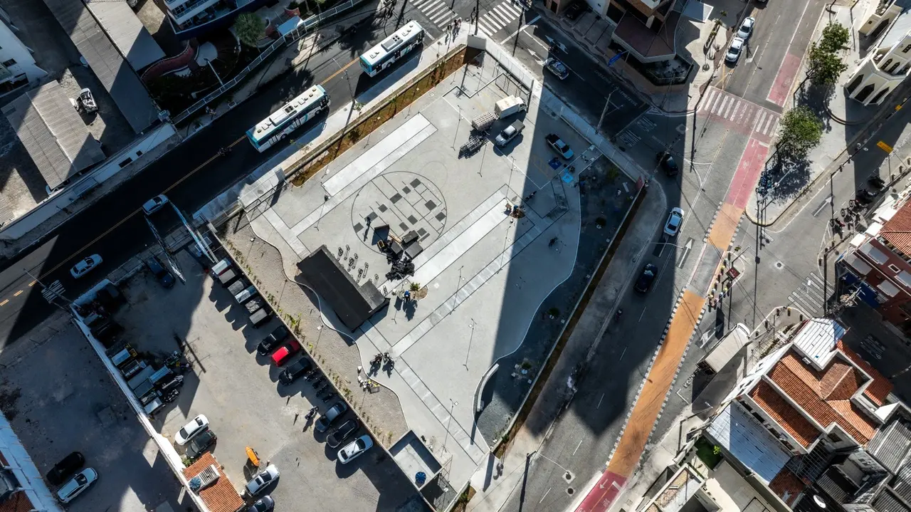 Vista aérea de uma praça urbana moderna (Praça Cultural São Pedro da UFC), com pavimento de concreto cinza e um padrão circular central. Ela está cercada por ruas movimentadas e estacionamentos, sob luz solar intensa, criando sombras fortes.