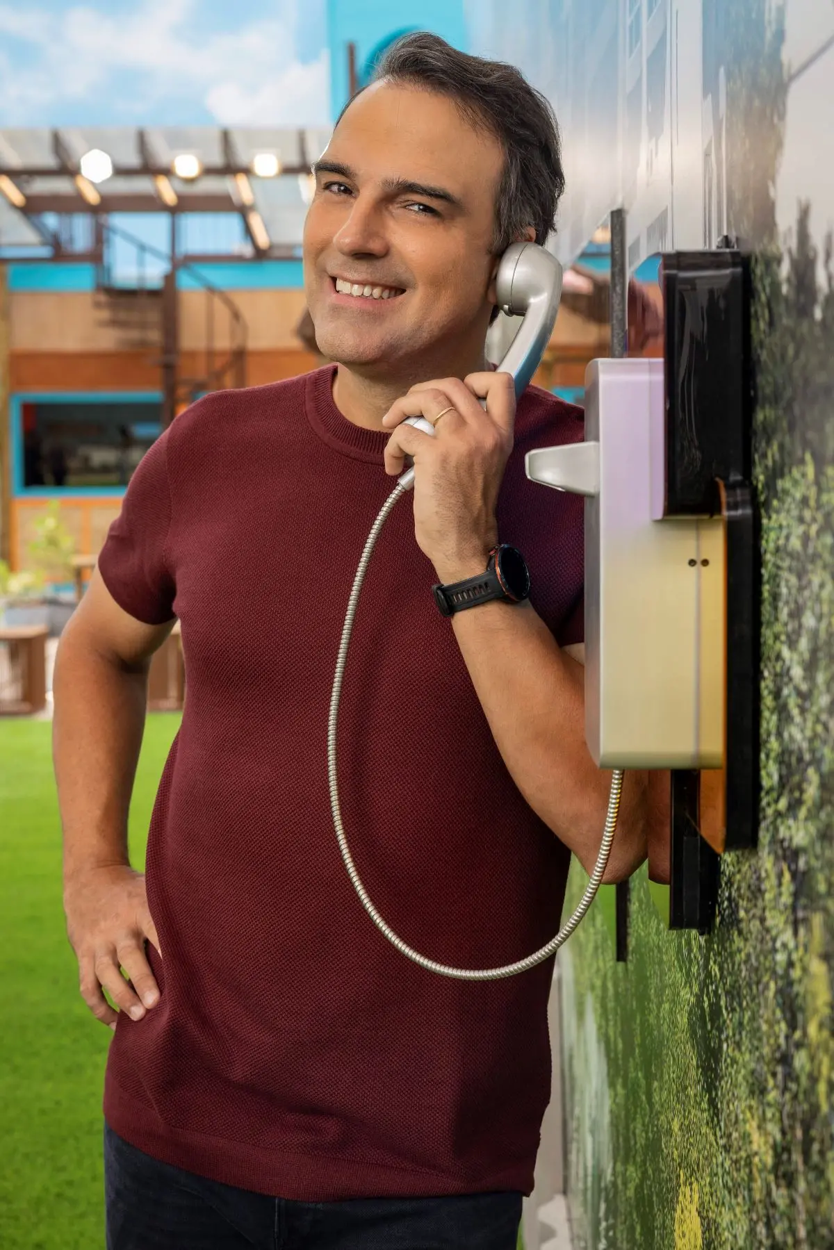 Pessoa usando uma camiseta de manga curta na cor vinho e um smartwatch preto, encostada em uma parede enquanto segura um telefone fixo com fio e design retrô. Ao fundo, área externa com gramado verde, estrutura de madeira e detalhes em azul sob céu parcialmente nublado.