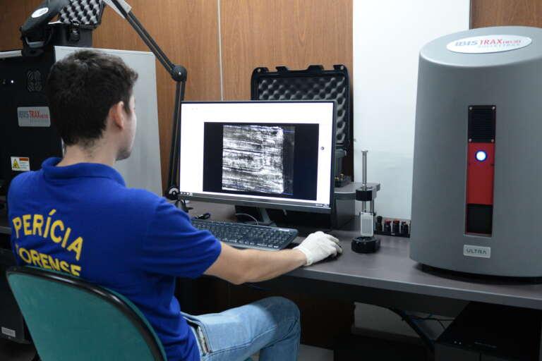 A imagem mostra um perito forense, de costas, blusa azul, olhando para um computador, onde ele realiza uma análise balística.