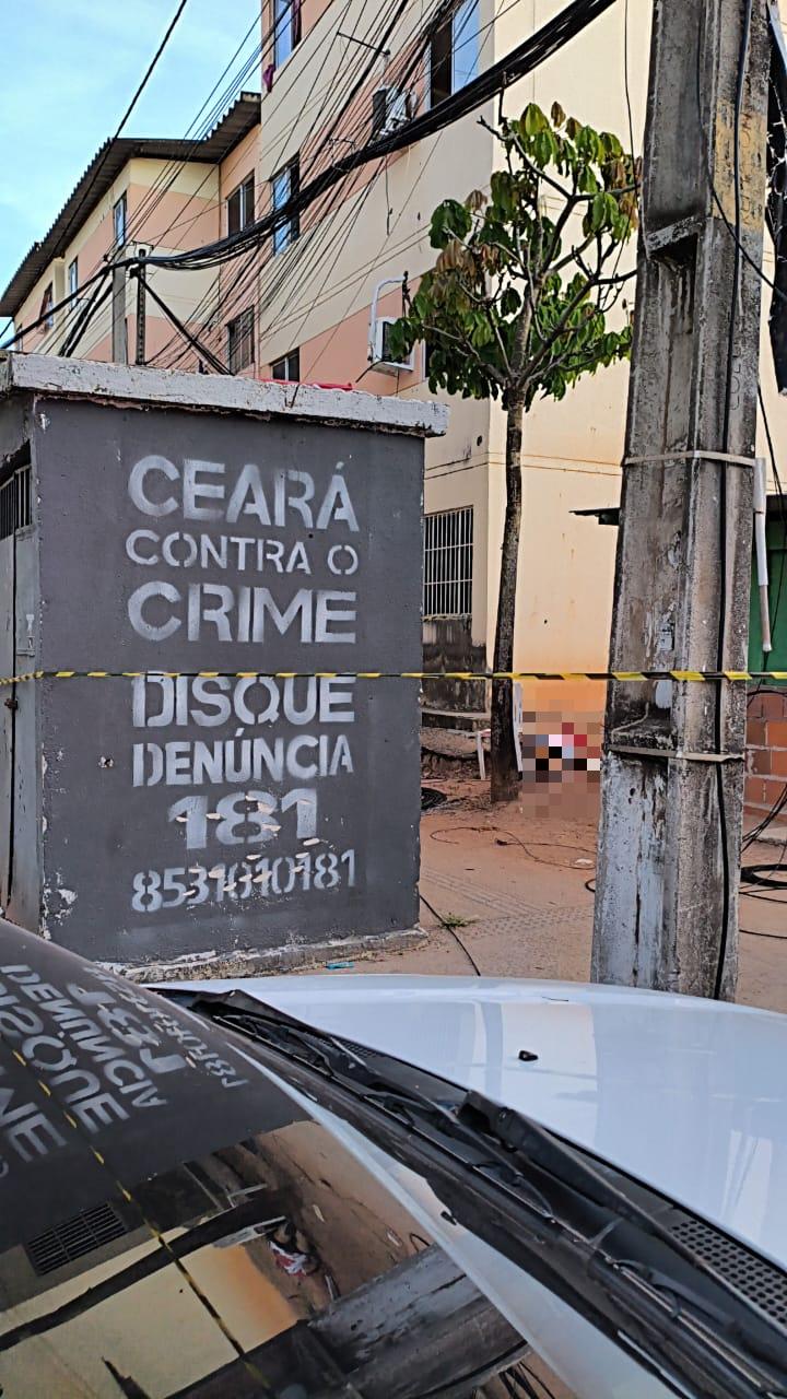 Guarita cinza com inscrição “Ceará contra o crime – Disque denúncia 181” e número de telefone, localizada em área urbana com prédios residenciais ao fundo, fios elétricos e veículo branco parcialmente visível na frente, cena delimitada por fita de isolamento policial.