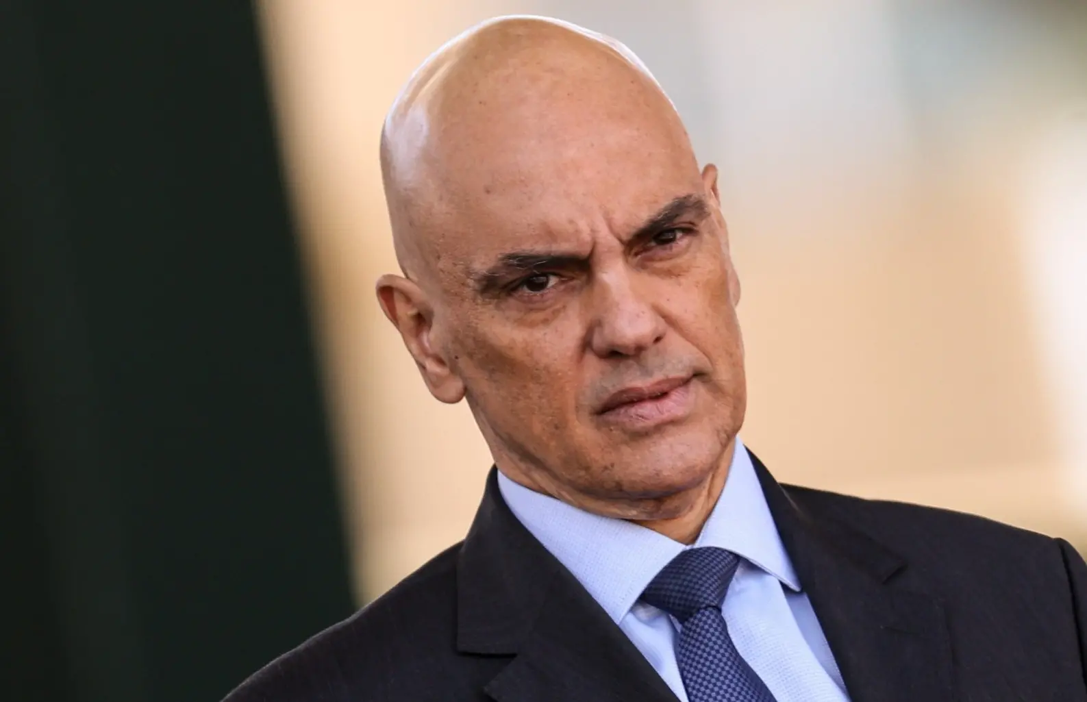Alexandre de Moraes é um homem de meia idade, careca e branco. Na foto, ele está com expressão séria e utiliza um terno preto com camisa social azul e gravata da mesma cor.