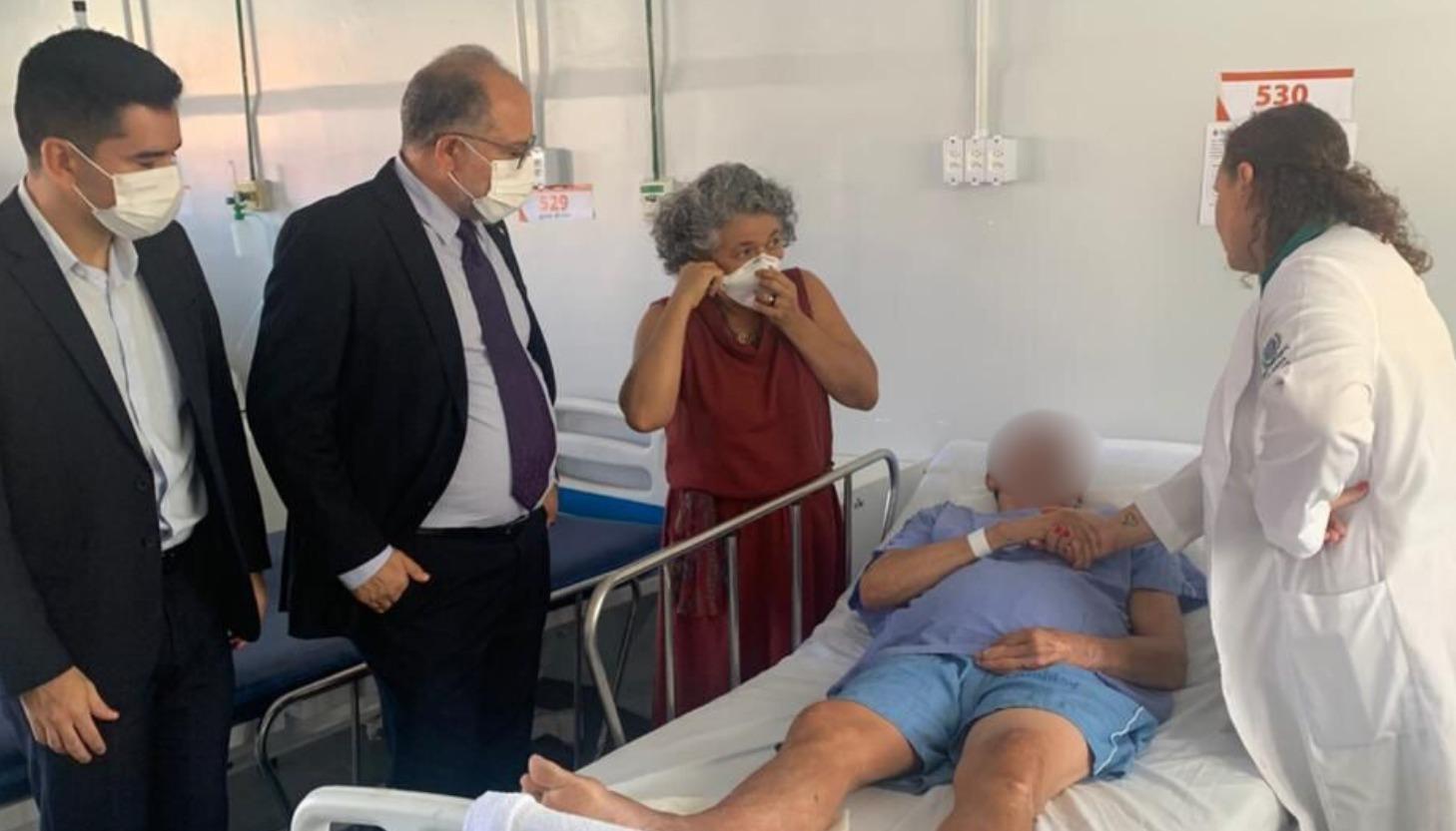Idoso deitado em uma cama é cumprimentado por uma mulher vestida de janelo. Do outro lado estão dois homens de paletó.