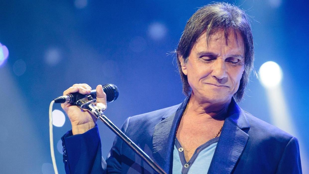 A imagem mostra o cantor Roberto Carlos, um homem idoso, de cor parda, cabelo grisalho e liso, com uma roupa azul, segurando um microfone.