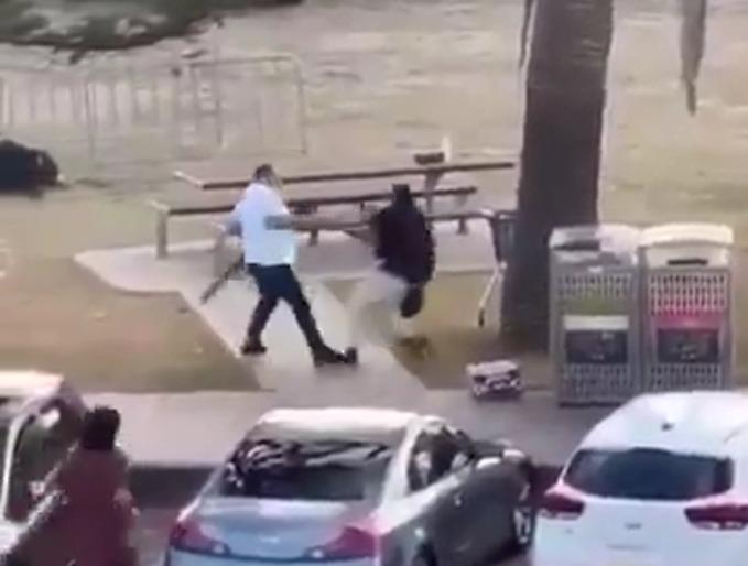 A imagem, print de um vídeo, mostra um homem com blusa branca tentando desarmar um homem com blusa preta, que portava uma arma de fogo longa.