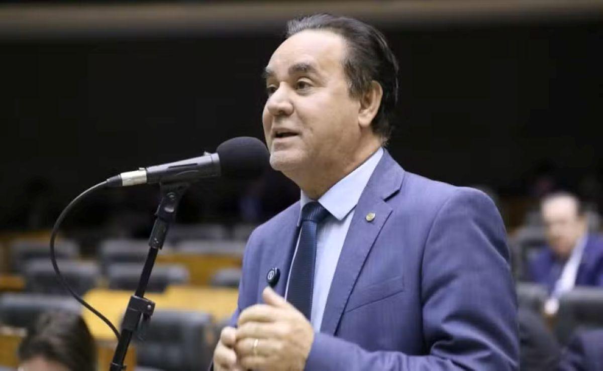 Político discursando em plenário parlamentar, usando terno e gravata, microfone à frente, ambiente legislativo com outros membros ao fundo.