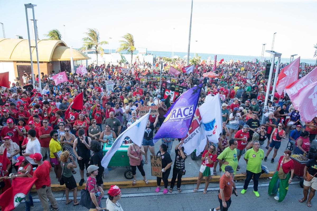 Multidão participa de manifestação ao ar livre em área urbana próxima ao mar, reunindo centenas de pessoas. A maioria veste camisetas vermelhas e carrega bandeiras, faixas e cartazes de movimentos sociais e sindicatos, com palavras de ordem e mensagens políticas. No primeiro plano, manifestantes seguram bandeiras brancas, roxas e vermelhas, enquanto outros observam ou conversam. Ao fundo, vê-se o calçadão, postes de iluminação, palmeiras e o oceano, indicando que o protesto ocorre durante o dia, em um espaço público amplo e movimentado.