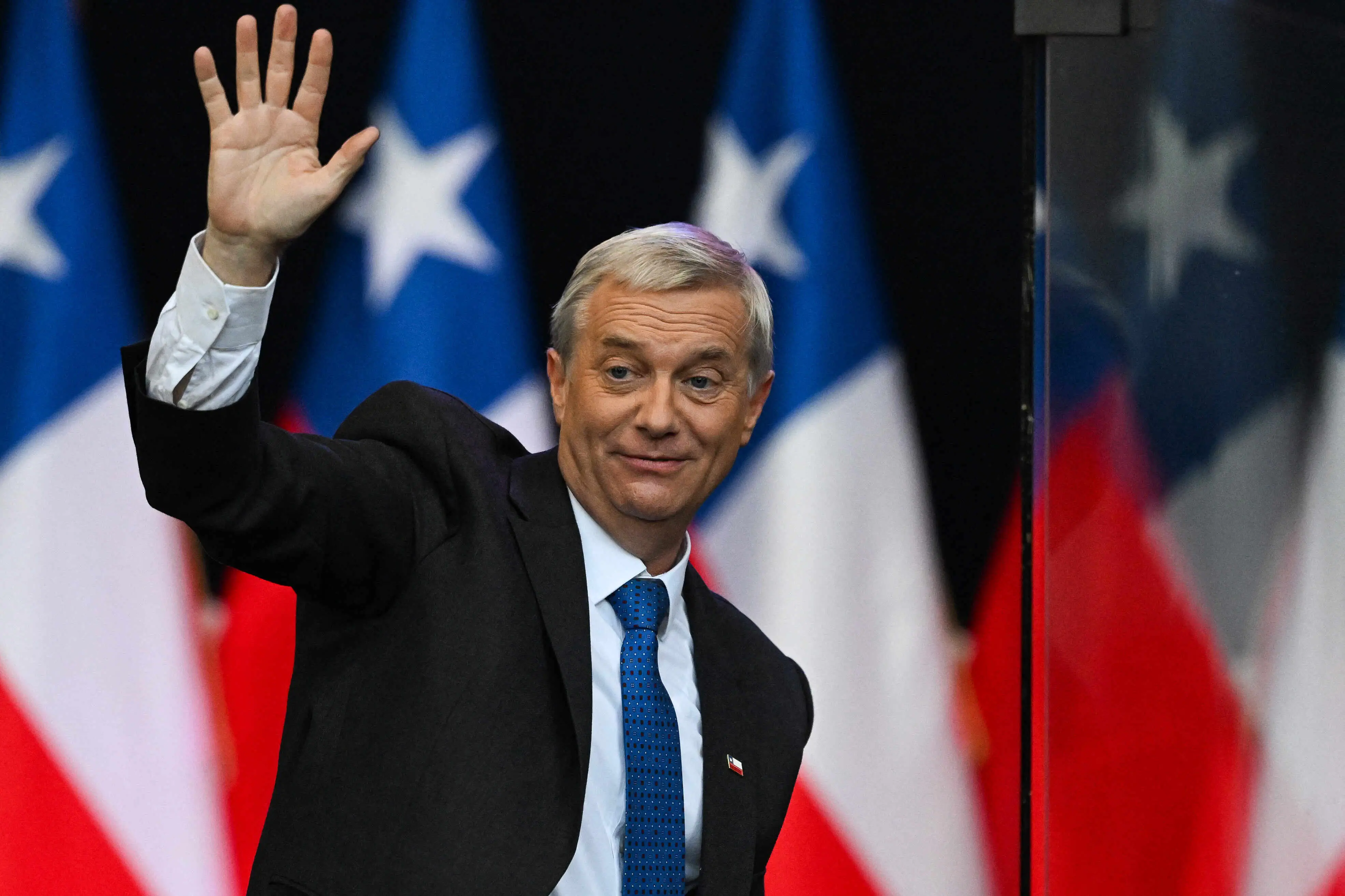 Homem de meia-idade, de cabelos grisalhos, usando terno escuro, camisa branca e gravata azul, acena com a mão direita enquanto sorri levemente. Ao fundo, aparecem várias bandeiras do Chile, desfocadas, em tons de azul, branco e vermelho.