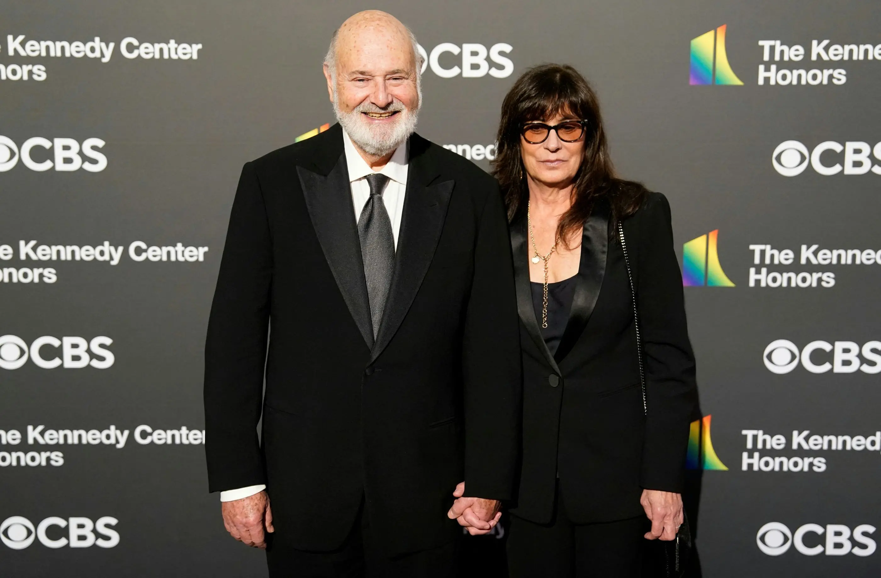 Rob Reiner e a esposa Michele diante de um painel com a marca da emissora CBS. eles vestem roupas pretas.