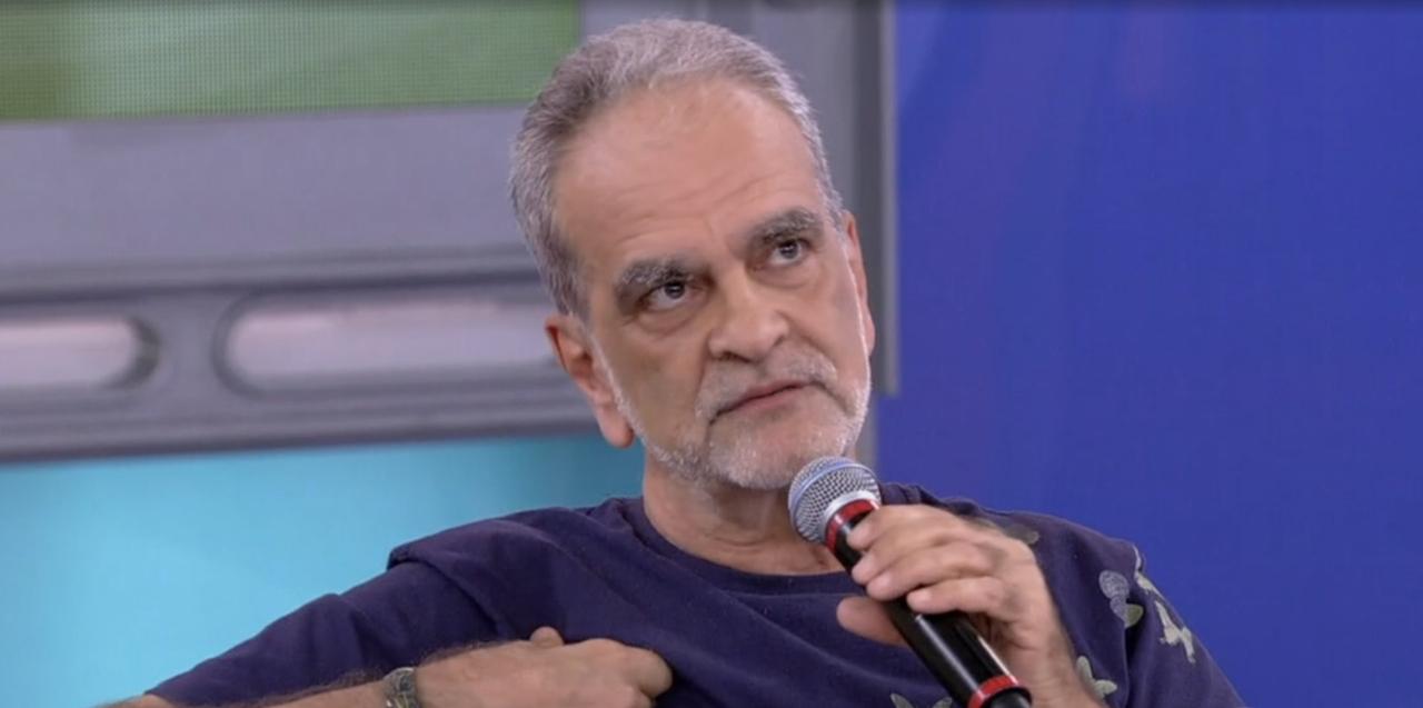 Homem pardo de barba rala branca e cabelos brancos, segurando microfone enquanto está sentado. Ele veste uma blusa azul marinho de manga semi longa.