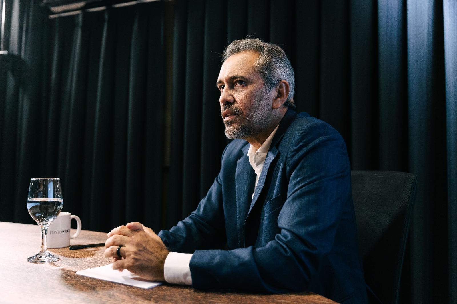 Elmano de Freitas de terno azul, sentado à mesa durante entrevista. Ele mantém as mãos apoiadas sobre a mesa, com um copo de água e uma caneca ao lado, diante de um fundo escuro com cortinas.