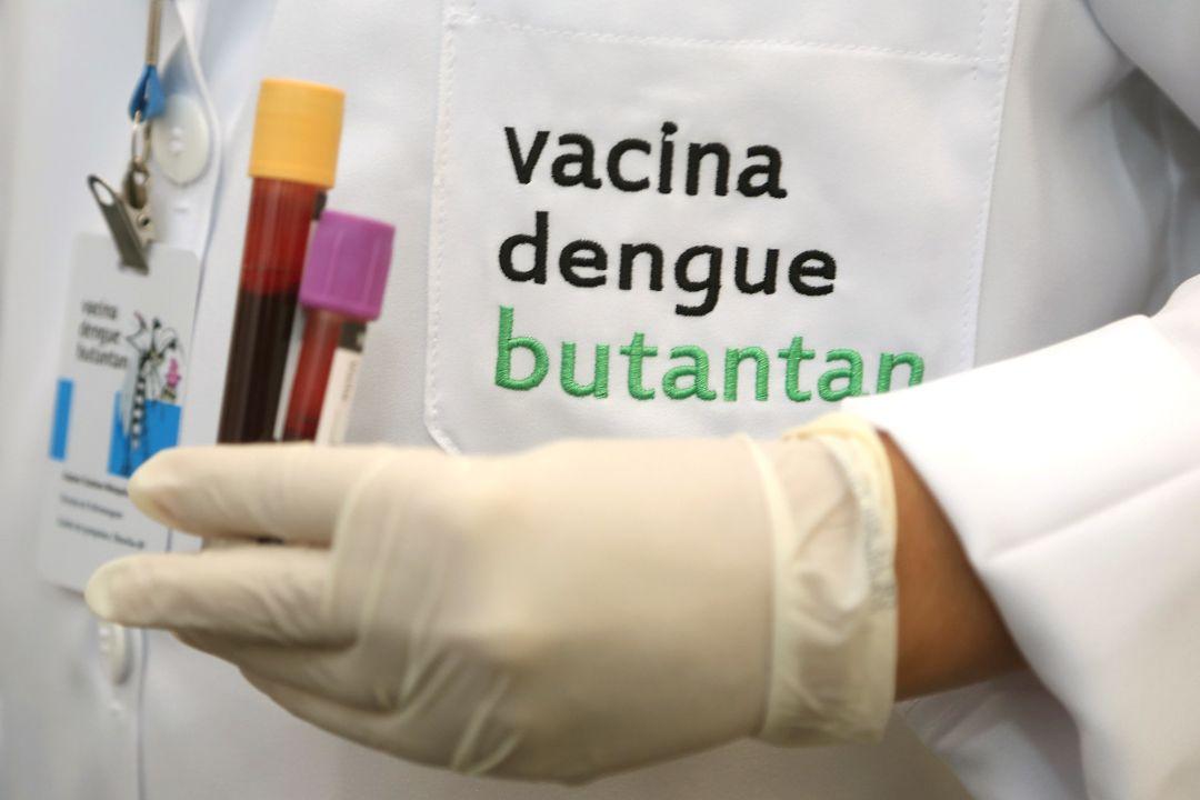 Profissional da saúde segura dois tubos de sangue com uma mão com luva. Ao lado, um bolso tem bordada a frase vacina dengue butantan.